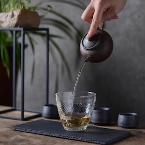 May include: A dark brown ceramic teapot pouring tea into a clear glass cup with a textured surface. The teapot has a round body and a small spout. The tea is a light amber color. The cup is sitting on a black slate serving tray.