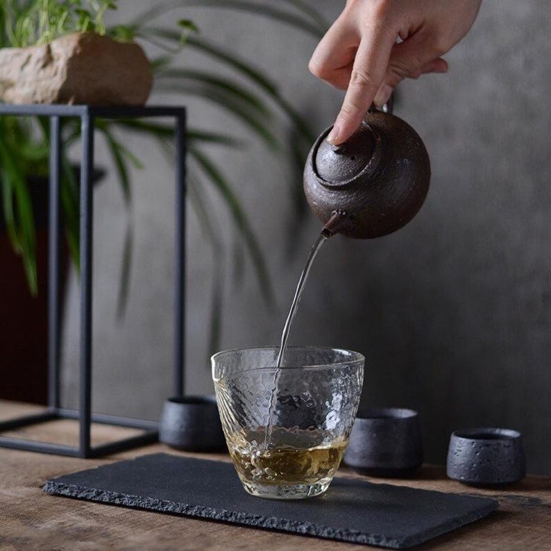May include: A dark brown ceramic teapot pouring tea into a clear glass cup with a textured surface. The teapot has a round body and a small spout. The tea is a light amber color. The cup is sitting on a black slate serving tray.