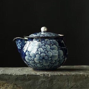 May include: A blue and white porcelain teapot with a floral design. The teapot has a lid and a spout. It is sitting on a gray surface.