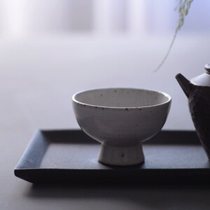 May include: A white ceramic bowl with a pedestal base sits on a dark grey rectangular tray. The bowl has a speckled texture and a rounded shape. A dark teapot is partially visible to the right.