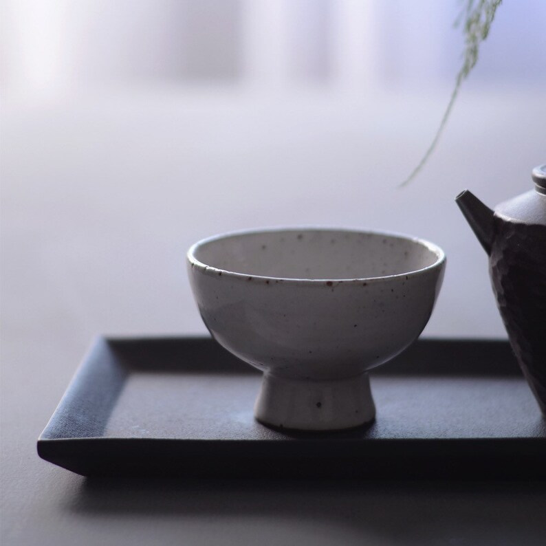May include: A white ceramic bowl with a pedestal base sits on a dark grey rectangular tray. The bowl has a speckled texture and a rounded shape. A dark teapot is partially visible to the right.