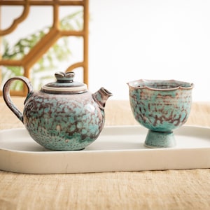 Puede incluir: Un juego de té de cerámica con un esmalte moteado en verde azulado y marrón. El juego incluye una tetera con tapa y asa, y una taza de té sobre una base de pedestal. El juego se exhibe en una bandeja ovalada blanca.