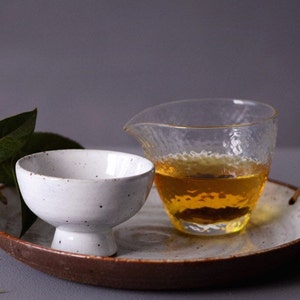 May include: A white ceramic tea bowl with a pedestal base and a speckled finish sits on a tray next to a clear glass pitcher filled with amber liquid. The tray has a brown rim and woven handles.