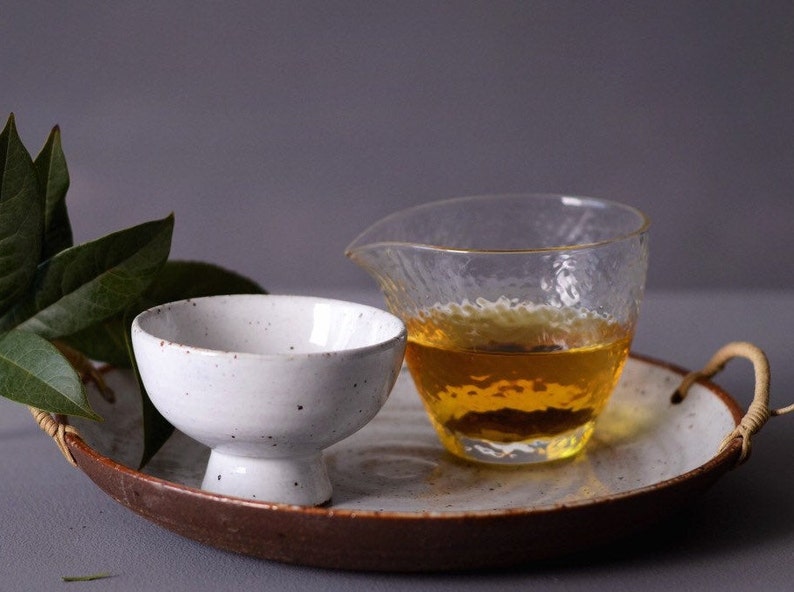 May include: A white ceramic tea bowl with a pedestal base and a speckled finish sits on a tray next to a clear glass pitcher filled with amber liquid. The tray has a brown rim and woven handles.