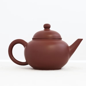 Op de afbeelding: Een kastanjebruine theepot met een afgeronde body, een gebogen handvat en een tuit. Het deksel heeft een kleine, ronde knop. De theepot is gemaakt van keramiek of porselein en staat op een witte ondergrond. Het ontwerp is eenvoudig en elegant.
