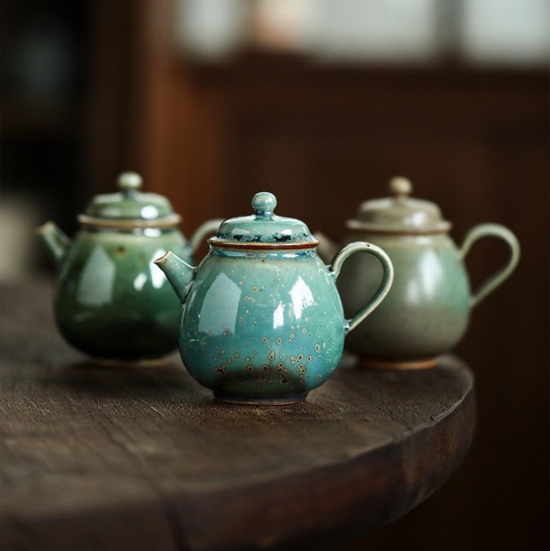 May include: Three ceramic teapots in shades of green and teal sit on a wooden surface. The teapots have rounded bodies, small spouts, and matching lids with small knobs. The teal teapot in the center has a speckled glaze.