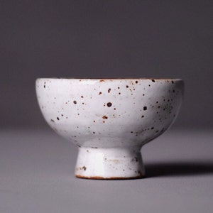 May include: A small, white ceramic bowl with a speckled design. The bowl has a rounded top and a short, pedestal base. The white surface is dotted with small, brown speckles, giving it a rustic appearance. The bowl is likely used for serving small portions.