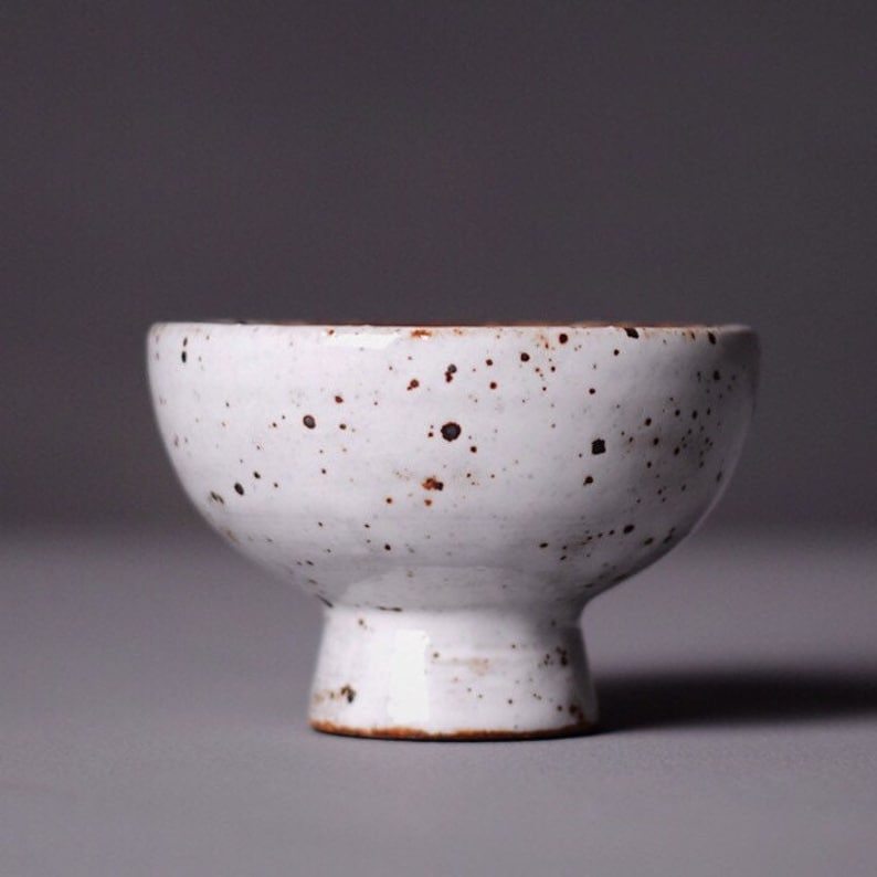 May include: A small, white ceramic bowl with a speckled design. The bowl has a rounded top and a short, pedestal base. The white surface is dotted with small, brown speckles, giving it a rustic appearance. The bowl is likely used for serving small portions.