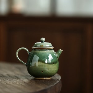 May include: A ceramic teapot with a rounded body and a curved handle. The teapot is glazed in shades of green, with a lid and a small spout. The teapot is sitting on a wooden surface.
