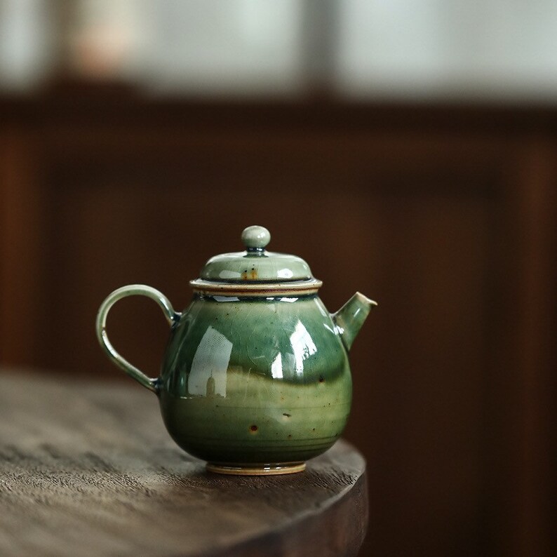 May include: A ceramic teapot with a rounded body and a curved handle. The teapot is glazed in shades of green, with a lid and a small spout. The teapot is sitting on a wooden surface.
