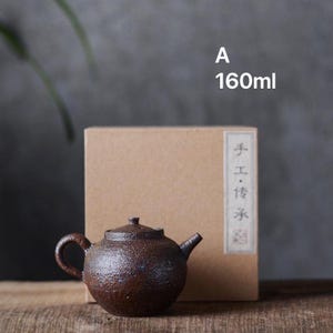 May include: A small, dark brown ceramic teapot with a lid and a spout. The teapot is sitting on a wooden surface in front of a brown cardboard box. The box has the text "手工&middot;传承" written on it. The text "A 160ml" is written in white on a gray background above the box.