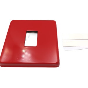 May include: A red metal light switch cover plate with a rectangular cutout for a light switch. The plate is designed to be mounted on a wall.