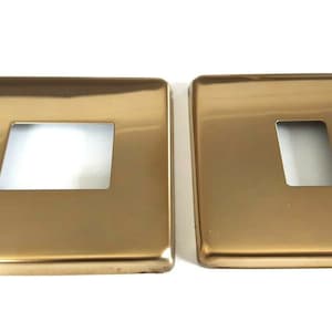 May include: Two gold-coloured metal switch plates with a rectangular cutout for a light switch.