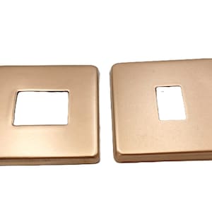 May include: Two square, gold-coloured light switch plates. Each plate has a rectangular opening in the centre. The plates are made of a smooth, metallic material. The plates are designed for electrical fixtures.