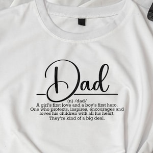 May include: White t-shirt featuring the word "Dad" in a decorative script. The shirt includes a definition: "A girl's first love and a boy's first hero. One who protects, inspires, encourages and loves his children with all his heart. They're kind of a big deal."
