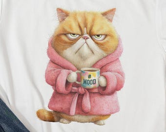May include: White t-shirt featuring a grumpy orange cat wearing a pink robe and holding a coffee mug with the word "MOOD". The cat has an annoyed expression. A black camera and other items are in the background.