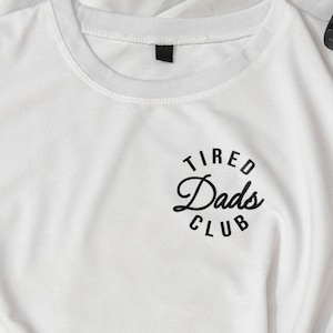 May include: A white t-shirt featuring the text "Tired Dads Club" in a black, circular design on the upper left chest. The shirt is made of a soft material and is knotted at the bottom. A camera and jeans are also visible.