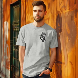 Pode incluir: Uma camiseta cinza com uma m&atilde;o de esqueleto preta fazendo um gesto de rock and roll com uma borboleta no dedo indicador. A m&atilde;o est&aacute; cercada por pequenas estrelas.