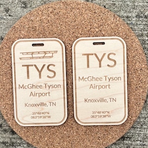 Op de afbeelding: Twee houten bagagelabels met de tekst "TYS McGhee Tyson Airport Knoxville, TN" en coördinaten. De labels zijn lichtbruin en liggen op een kurken onderzetter.