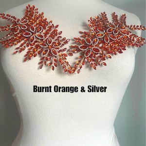 K&ouml;nnte beinhalten: Zwei dekorative Blumenapplikationen in gebranntem Orange und Silber. Jede Applikation hat ein zentrales Blumendesign, das von kleineren, blattf&ouml;rmigen Verzierungen umgeben ist. Der Text "Burnt Orange & Silver" ist darunter abgebildet.