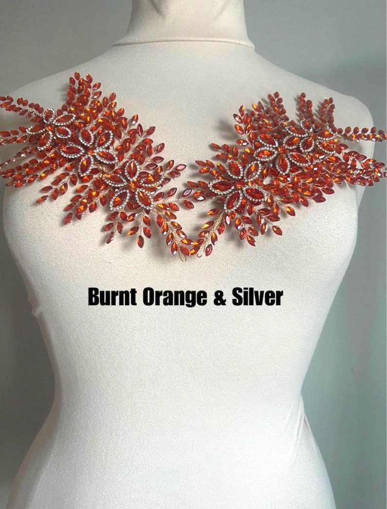 K&ouml;nnte beinhalten: Zwei dekorative Blumenapplikationen in gebranntem Orange und Silber. Jede Applikation hat ein zentrales Blumendesign, das von kleineren, blattf&ouml;rmigen Verzierungen umgeben ist. Der Text "Burnt Orange & Silver" ist darunter abgebildet.