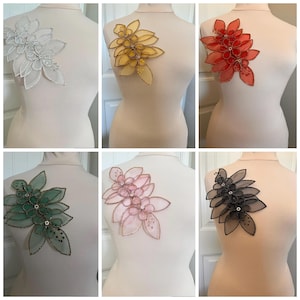 Puede incluir: Colección de seis apliques florales en blanco, amarillo, rojo, verde, rosa y negro. Cada aplique, fijado a un maniquí, presenta un diseño de pétalos superpuestos con detalles de cuentas, ideal para adornar prendas.