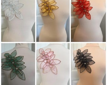 May include: A collection of six floral appliques in various colors, including white, yellow, red, green, pink, and black. Each applique is attached to a mannequin and features a layered petal design with beaded accents, suitable for embellishing garments.