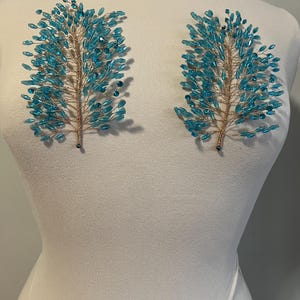 May include: Two teal beaded hair accessories in a tree branch design. The branches are made of gold wire and are adorned with numerous teal-colored beads. The accessories are displayed on a white mannequin.