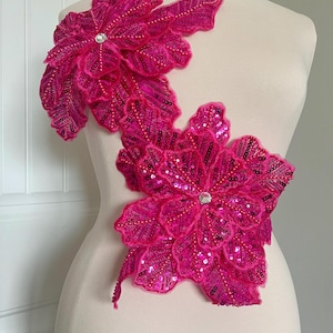 May include: Two large pink fabric flowers with sequins and beads. The flowers are layered on top of each other and have a rhinestone in the centre.