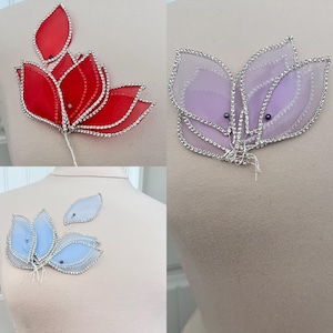 May include: Three floral brooches in red, lavender, and light blue. Each brooch features delicate, translucent petals outlined with silver rhinestones. The red brooch has multiple petals, while the lavender and blue brooches have fewer. The brooches are attached to a wire stem.