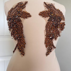 May include: Two ornate, symmetrical appliques with a leaf-like design. The appliques feature a complex arrangement of brown and amber-colored beads and sequins, attached to a sheer, flesh-toned backing. The embellishments are likely for formal wear.