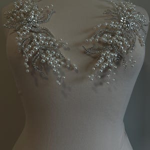 May include: Two silver and pearl beaded shoulder embellishments. Each features a central cluster of clear rhinestones surrounded by delicate silver wire branches adorned with small, round pearls. The embellishments are displayed on a mannequin.