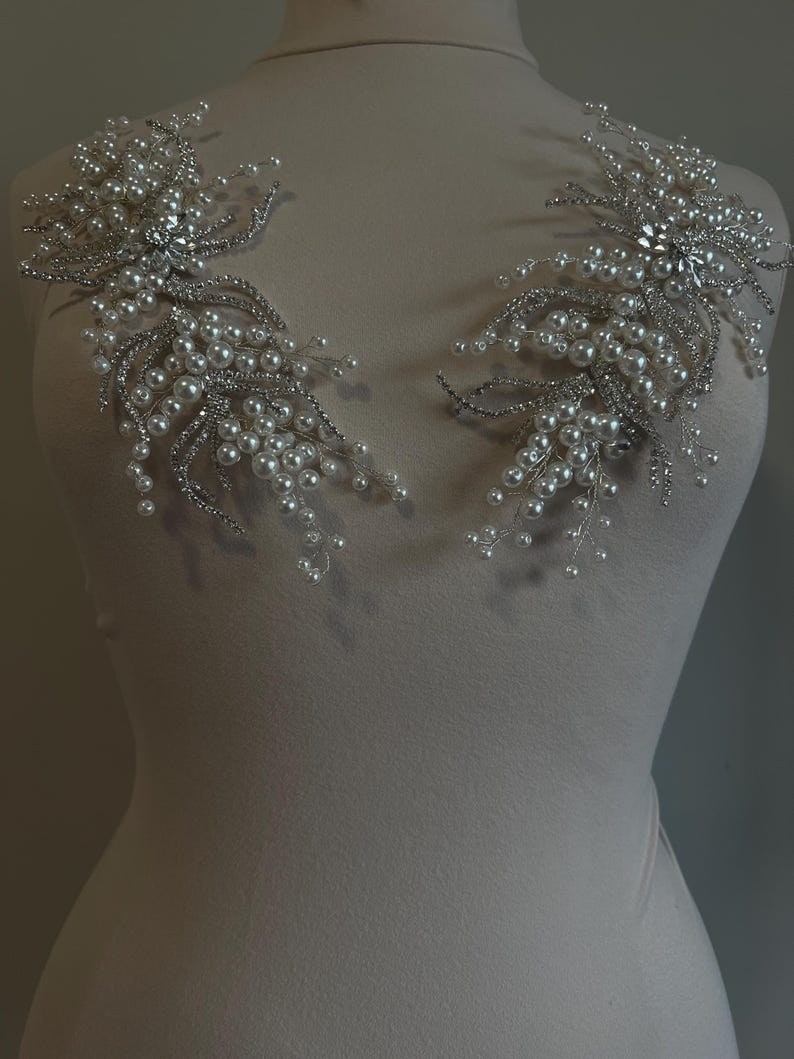 May include: Two silver and pearl beaded shoulder embellishments. Each features a central cluster of clear rhinestones surrounded by delicate silver wire branches adorned with small, round pearls. The embellishments are displayed on a mannequin.