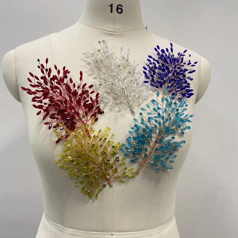 May include: Five beaded floral embellishments in red, white, blue, turquoise, and yellow-green are displayed on a mannequin. Each sprig-like design features small, sparkling beads attached to thin, wire stems. The mannequin is a neutral beige color.