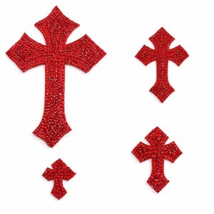 May include: Four red rhinestone crosses of varying sizes are arranged on a white background. The crosses have a detailed, textured appearance, with each facet reflecting light. The design is symmetrical, with the largest cross at the top.
