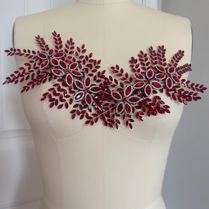 K&ouml;nnte beinhalten: Dekorative, rote Strass-Applikation mit floralem und Bl&auml;tter-Design. Die Applikation besteht aus zahlreichen roten Strasssteinen und silberfarbenem Metall, die in einem symmetrischen Muster angeordnet sind. Das Design wird auf einer Schaufensterpuppe pr&auml;sentiert.
