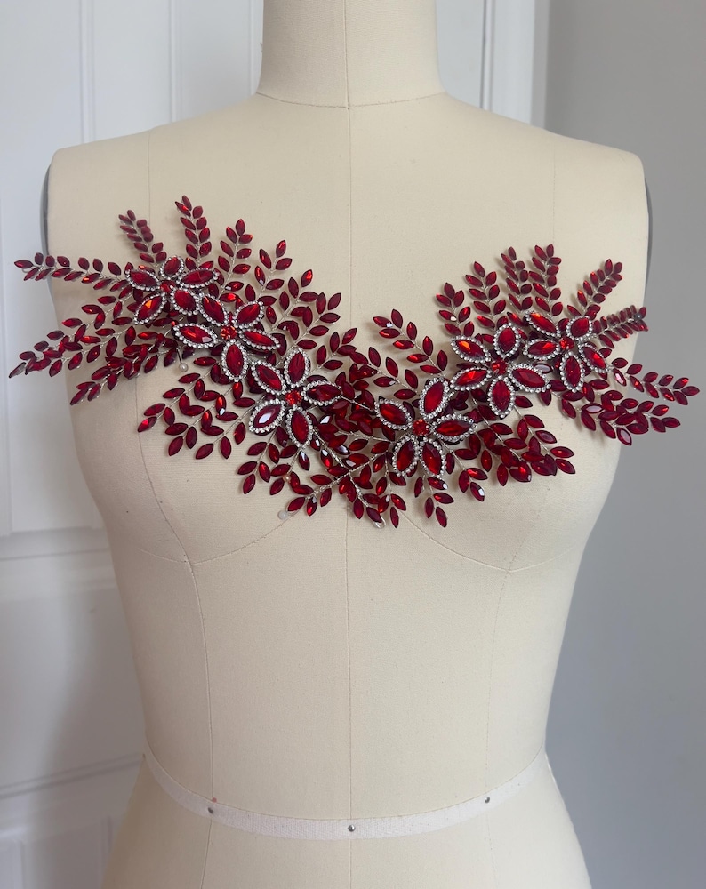 K&ouml;nnte beinhalten: Dekorative, rote Strass-Applikation mit floralem und Bl&auml;tter-Design. Die Applikation besteht aus zahlreichen roten Strasssteinen und silberfarbenem Metall, die in einem symmetrischen Muster angeordnet sind. Das Design wird auf einer Schaufensterpuppe pr&auml;sentiert.