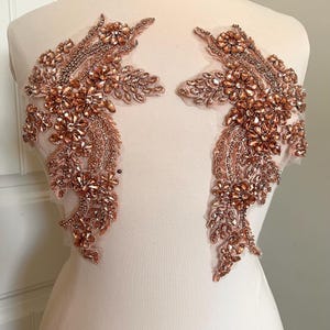 May include: Two copper-coloured, beaded floral appliques. Each applique features a detailed design of flowers and leaves, with a mix of matte and shiny beads. The appliques are set on a sheer, nude-coloured backing, creating a delicate and elegant look.