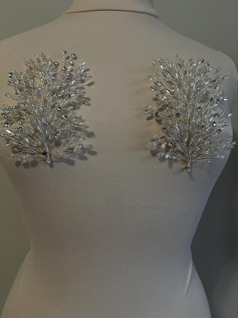 May include: Two silver-toned, crystal-embellished floral hair accessories. Each piece features a delicate wire framework adorned with clear, sparkling crystals, arranged in a branch-like design. The accessories are displayed on a mannequin, suggesting bridal or formal wear.