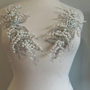 May include: Two silver and pearl beaded floral appliques. Each applique features clusters of white pearls and silver-toned wire branches, with a central cluster of clear rhinestones. The appliques are displayed on a mannequin.