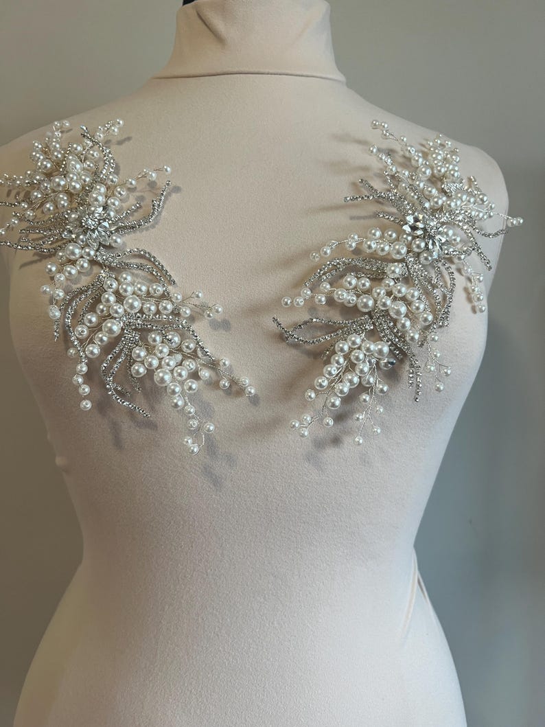 May include: Two silver and pearl beaded floral appliques. Each applique features clusters of white pearls and silver-toned wire branches, with a central cluster of clear rhinestones. The appliques are displayed on a mannequin.