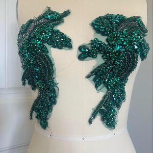 May include: Two green beaded appliques with a floral design. The appliques are on a white fabric background.