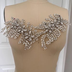 K&ouml;nnte beinhalten: Silberne und transparente Kristallapplikation mit floralem Design und mehreren Kristallakzenten. Die Applikation wird auf einer Schneiderpuppe pr&auml;sentiert und eignet sich f&uuml;r Braut- oder Abendmode.