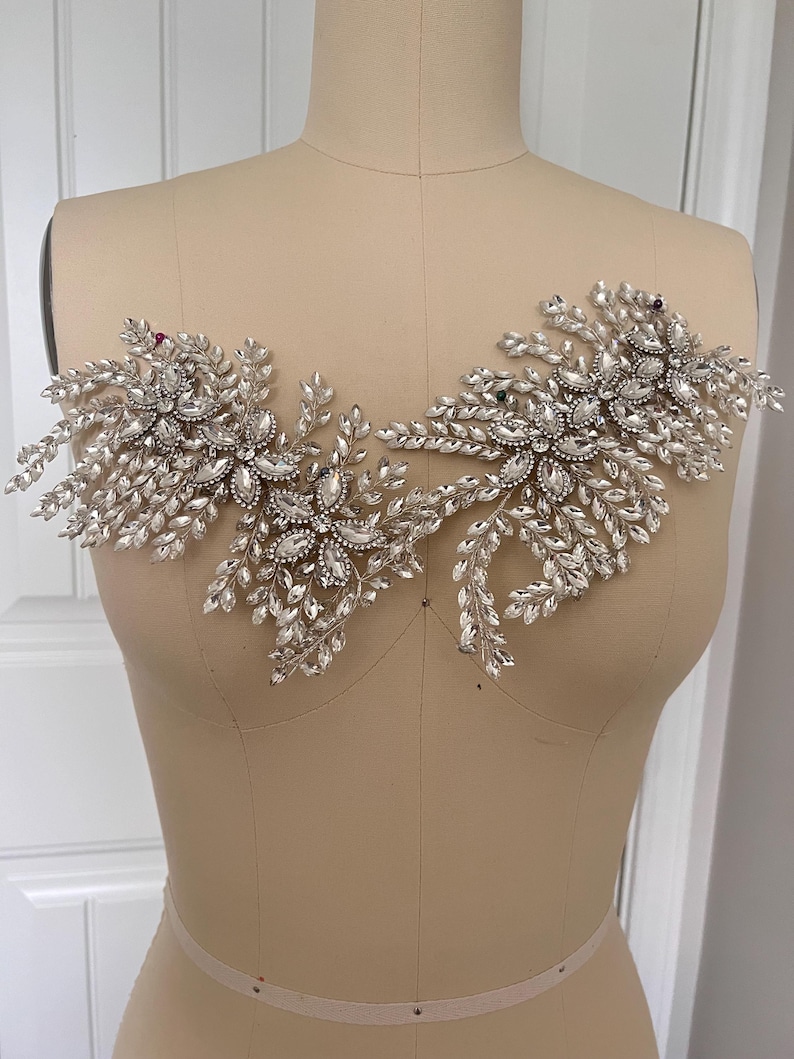 K&ouml;nnte beinhalten: Silberne und transparente Kristallapplikation mit floralem Design und mehreren Kristallakzenten. Die Applikation wird auf einer Schneiderpuppe pr&auml;sentiert und eignet sich f&uuml;r Braut- oder Abendmode.