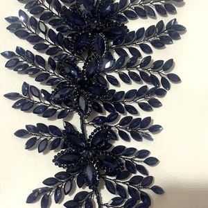 K&ouml;nnte beinhalten: Dunkelblaues Haaraccessoire mit floralem und Bl&auml;tter-Design. Es zeigt Gruppen dunkelblauer Juwelen und blattf&ouml;rmige Akzente, alle auf einem schwarzen Drahtrahmen. Das Design ist symmetrisch und elegant, ideal f&uuml;r besondere Anl&auml;sse.