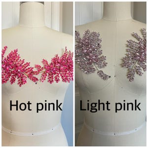 K&ouml;nnte beinhalten: Zwei dekorative Blumenapplikationen, eine in Pink und die andere in Hellrosa, werden auf Schneiderpuppen gezeigt. Die pinkfarbene Applikation befindet sich links, die hellrosa Applikation rechts. Text darunter zeigt die Farbe an.