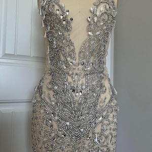 May include: A silver beaded and sequined embellishment for a dress or garment. The embellishment is in a floral pattern with a sheer fabric backing.