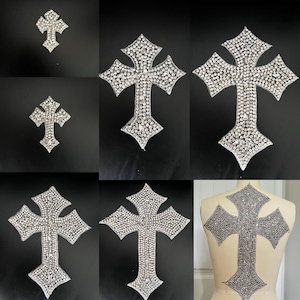 May include: A collection of rhinestone cross appliques in various sizes. The crosses are silver and feature a detailed pattern of sparkling rhinestones. One applique is shown on a mannequin.