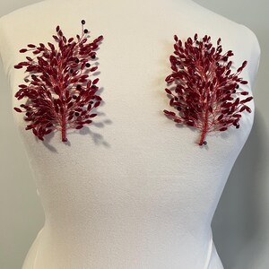 May include: Two red beaded floral embellishments, each with a branch-like design, are displayed on a white mannequin. The embellishments feature small, dark beads interspersed among the red beads, creating a textured appearance. These could be used for bridal or formal wear.