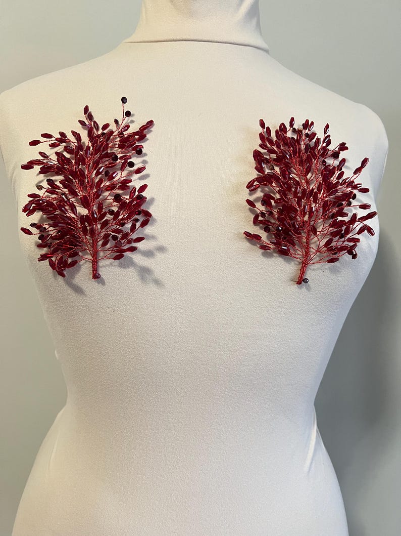 May include: Two red beaded floral embellishments, each with a branch-like design, are displayed on a white mannequin. The embellishments feature small, dark beads interspersed among the red beads, creating a textured appearance. These could be used for bridal or formal wear.